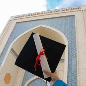A graduate holding up a degree diploma and graduation cap in front of an international university campus.