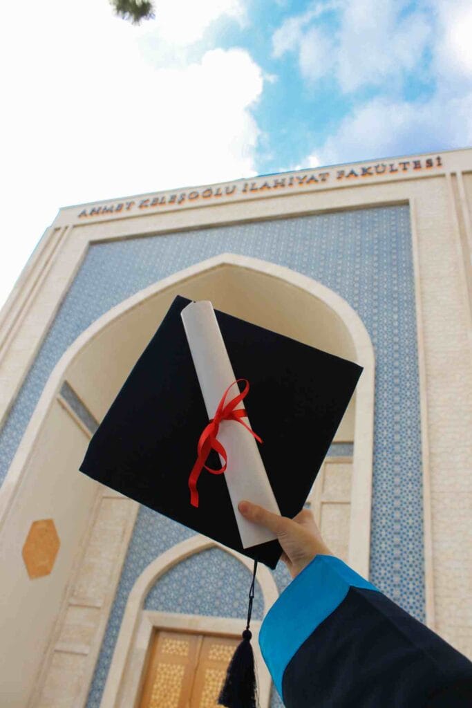 A graduate holding up a degree diploma and graduation cap in front of an international university campus.